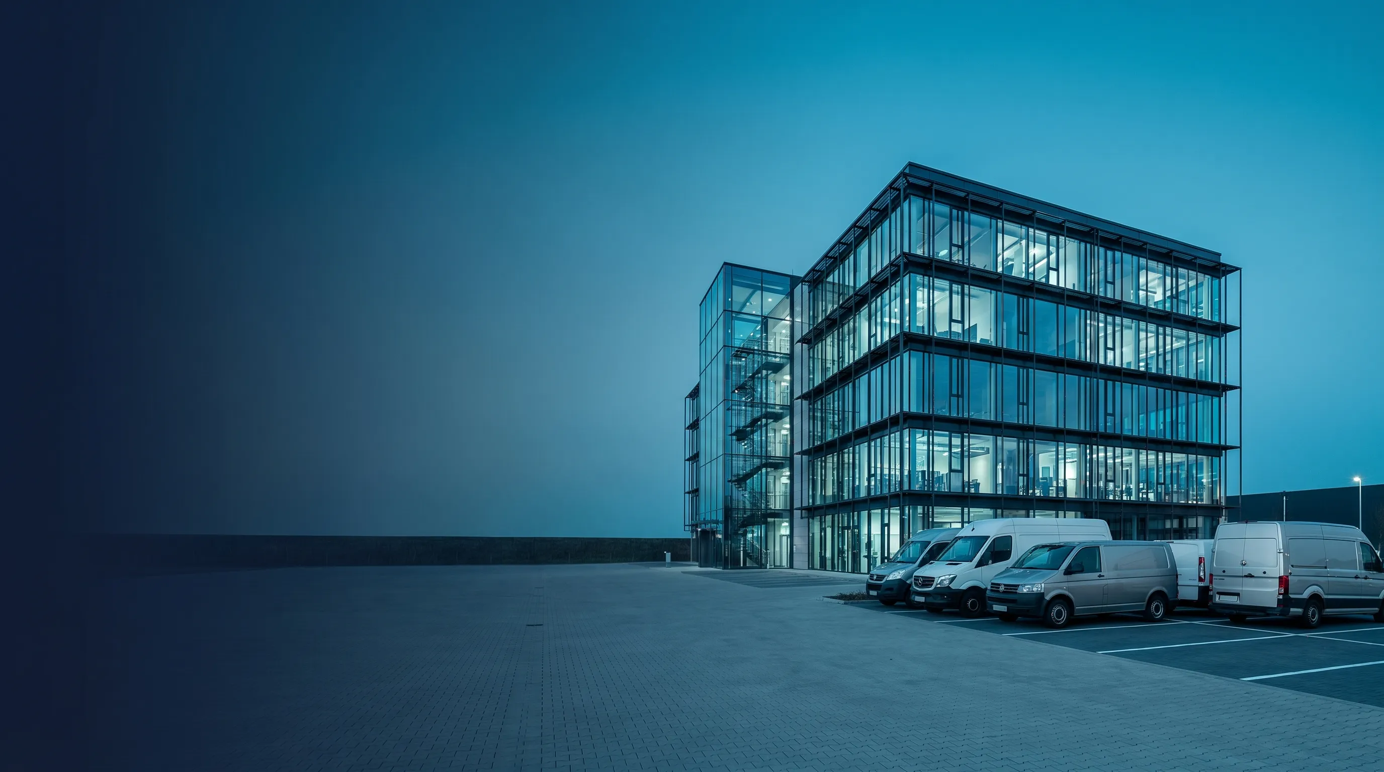 Modernes Bürogebäude mit Servicefahrzeugen auf dem Parkplatz in der blauen Stunde, freie Fläche links für den Hero-Text